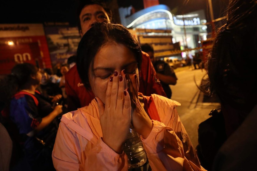 A woman standing on the street near a shopping mall in Thailand covers her face with her hands.