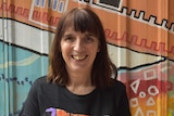 A smiling woman with brown hair smiles as she stands in front of a colourful wall.