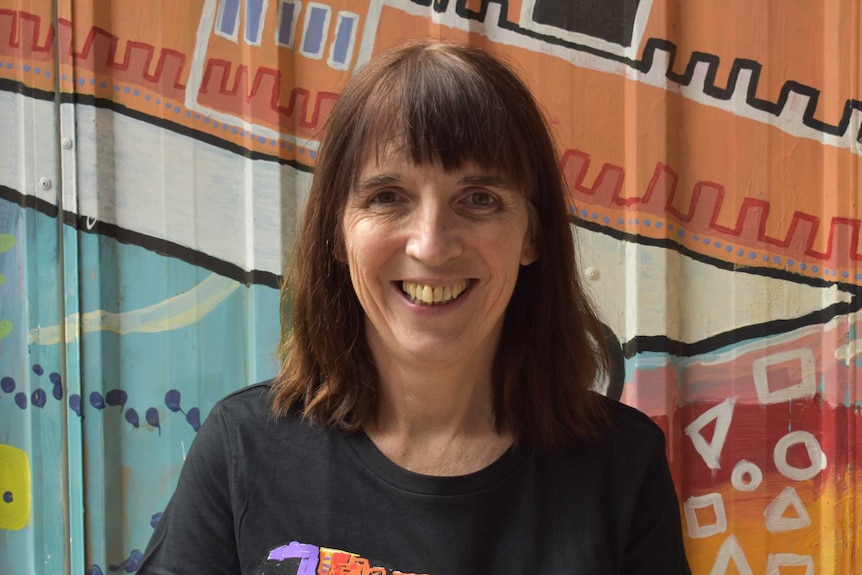 A smiling woman with brown hair smiles as she stands in front of a colourful wall.