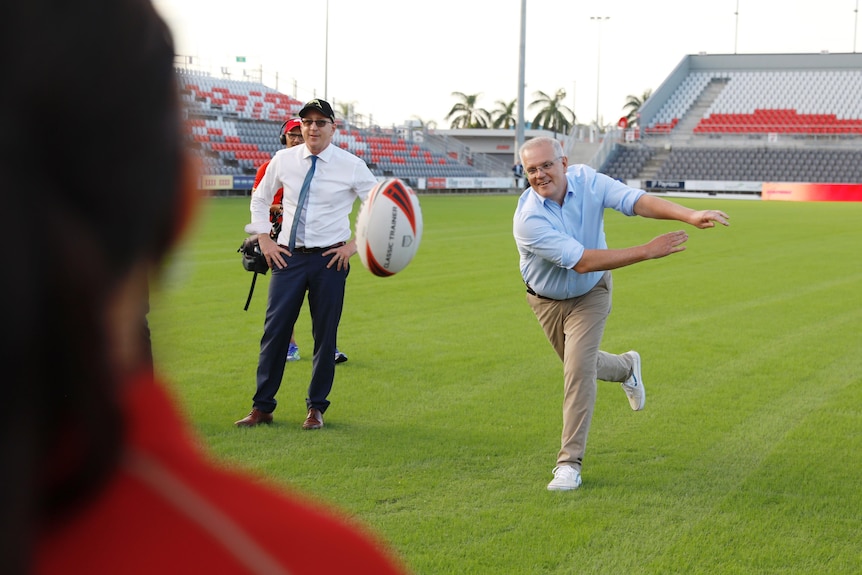 a man stands with his hands on his hips as Scott Morrison throws a rugby league ball toward a person in the foreground