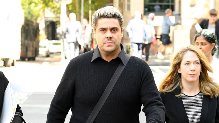 A tall man with blonde hair wearing black slacks, sweater and shirt walks along a street flanked by two women in business suits.