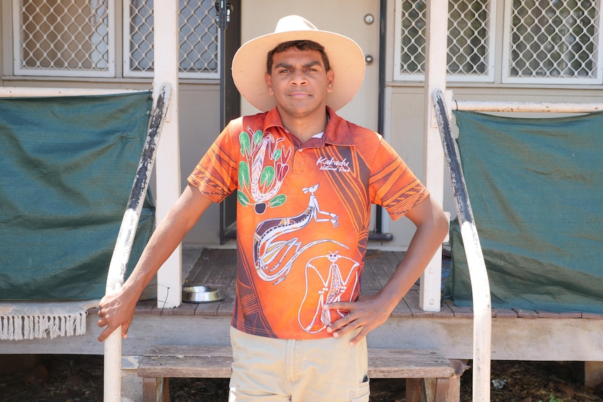 A young Indigenous man with a wide brimmed hat and an orange shirt looks straight at the camera