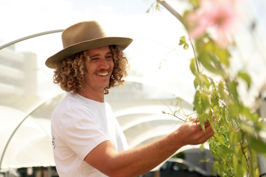 Jared wears a white shirt and brown wide brimmed hat and smiles as he checks a plant.