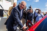 Scott Morrison refilling a red electric vehicle with a a crowd of people behind him.