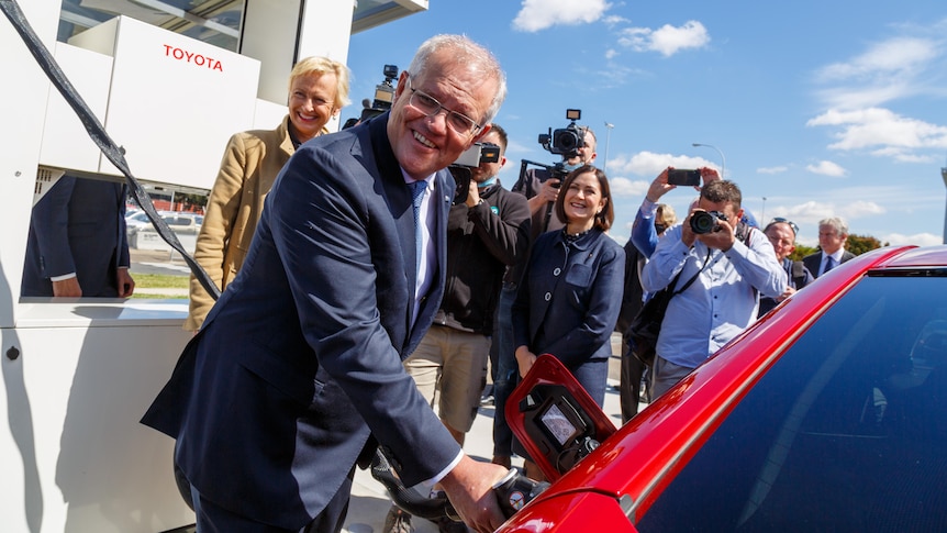 Scott Morrison refilling a red electric vehicle with a a crowd of people behind him.