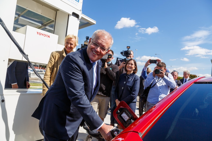 Scott Morrison refilling a red electric vehicle with a a crowd of people behind him.
