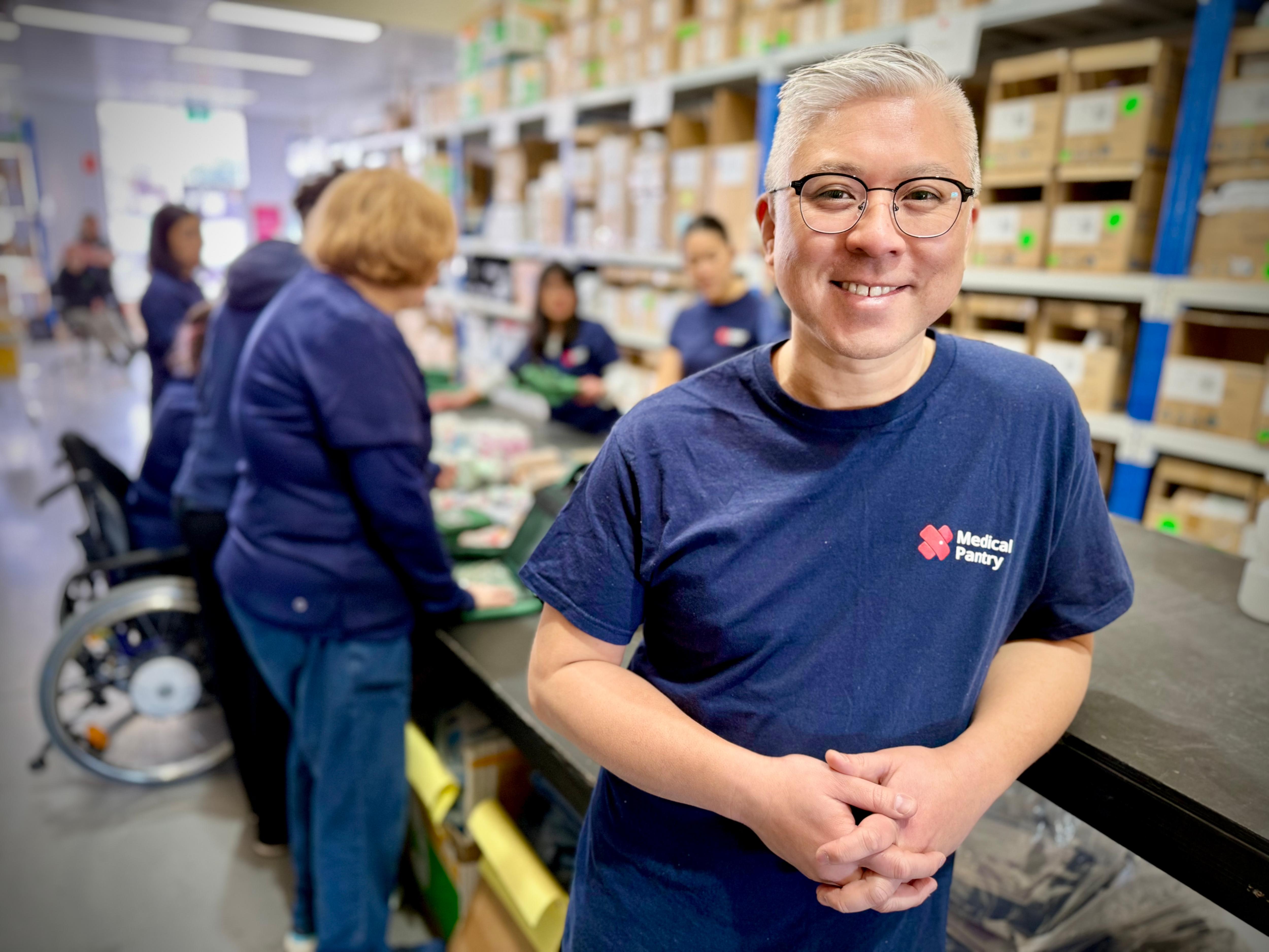 Um homem de cabelos brancos e óculos sorri para a câmera. Ele está em um armazém cheio de suprimentos médicos. 