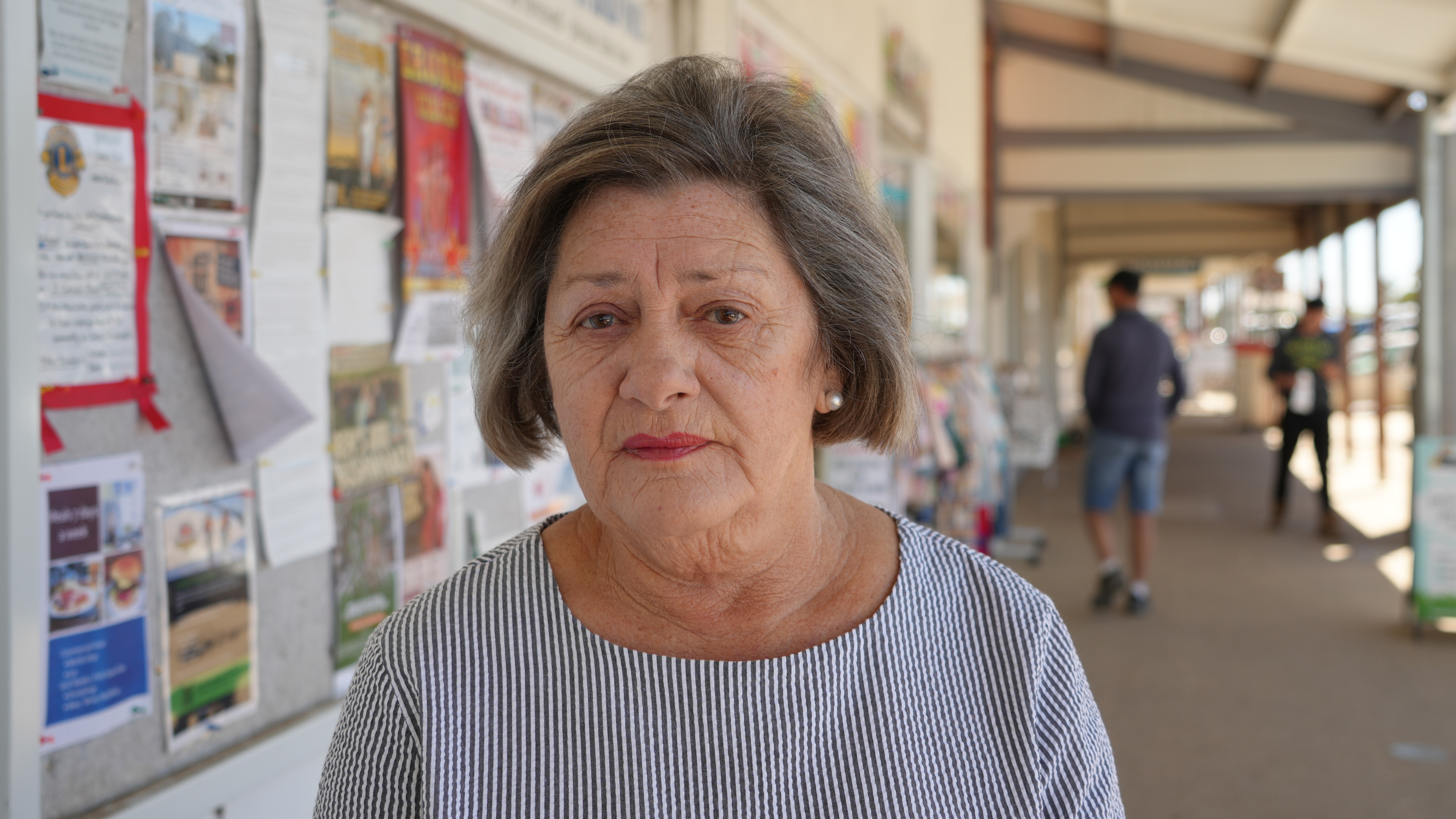 Elderly woman with short grey bob and red lipstick looks seriosuly at camera, standing in regional town street, outside shops