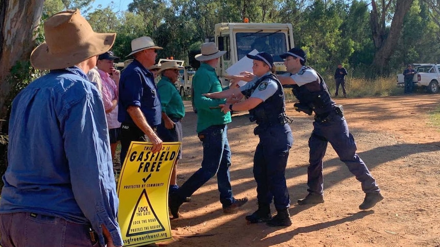 Five men and women stand in shirts and jeans as a police officer has their arms stretched to hold back one protester.