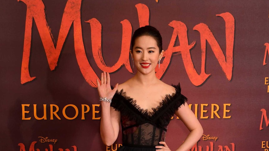 Chinese actress wearing a black ball gown stands in front of Disney's Mulan branded backdrop.