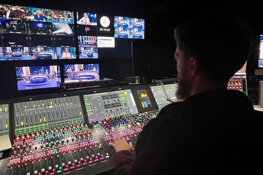 Man sitting at audio desk looking at TV monitors.