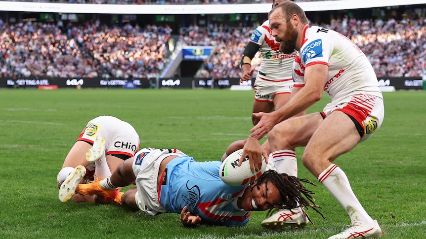 NRL player Dominic Young on the ground, reaching out wit the ball to the try line, with a defender holding onto his foot