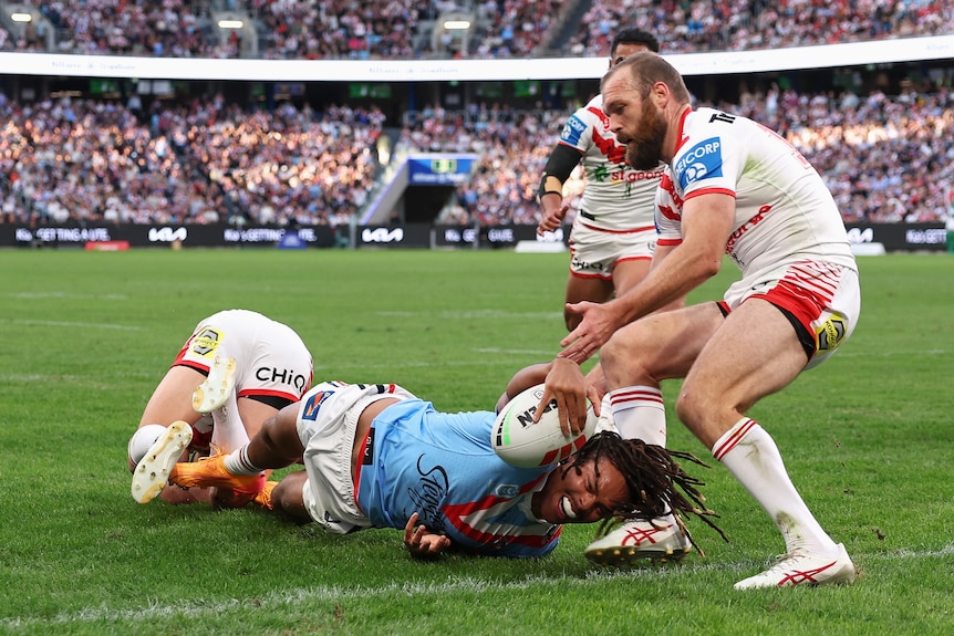 NRL player Dominic Young on the ground, reaching out wit the ball to the try line, with a defender holding onto his foot