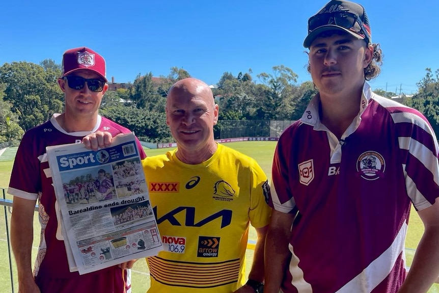 Three men smile for the camera. One his holding up a newspaper.