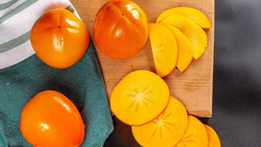 Sliced persimmons spread on and off chopping board next to three whole persimmons on a tea towel