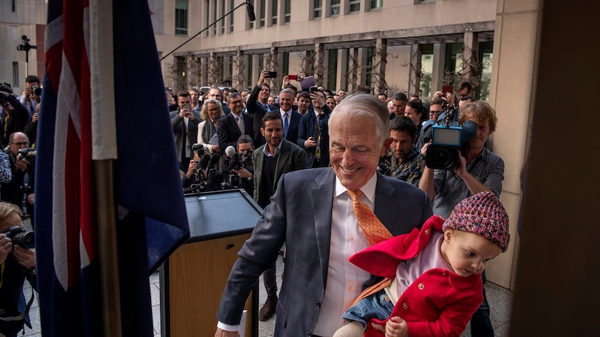 Smiling Malcolm Turnbull walks away away from the lectern, holding a child, with a large media contingent in the background