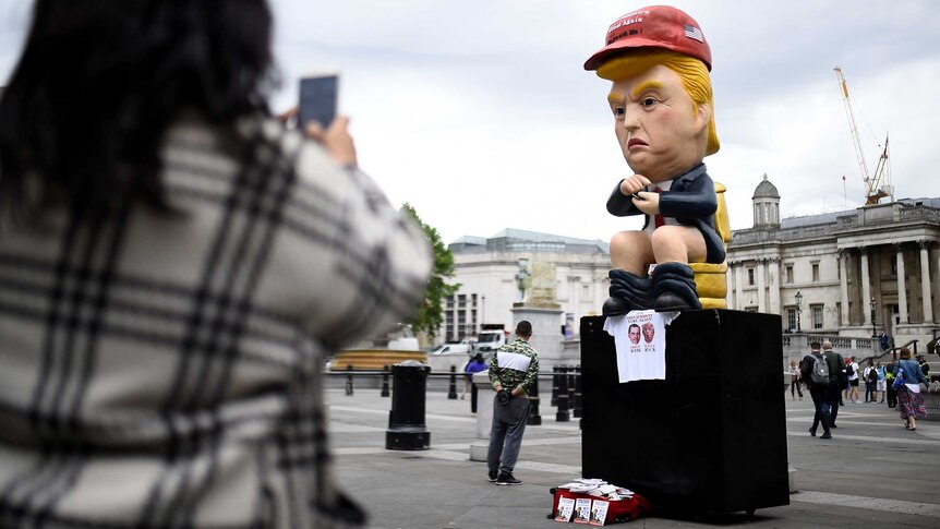 A person takes a photo of a cartoonish statue of Donald Trump, pictured sitting with pants around his ankles looking at a phone.