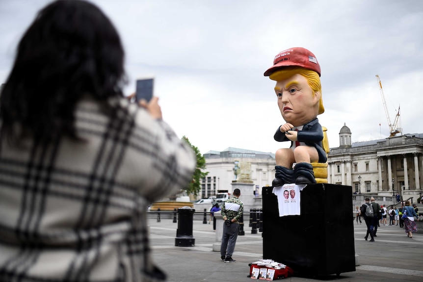 A person takes a photo of a cartoonish statue of Donald Trump, pictured sitting with pants around his ankles looking at a phone.