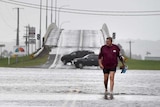 A man walks through ankle deep floodwaters, with cars and a bridge in the background.