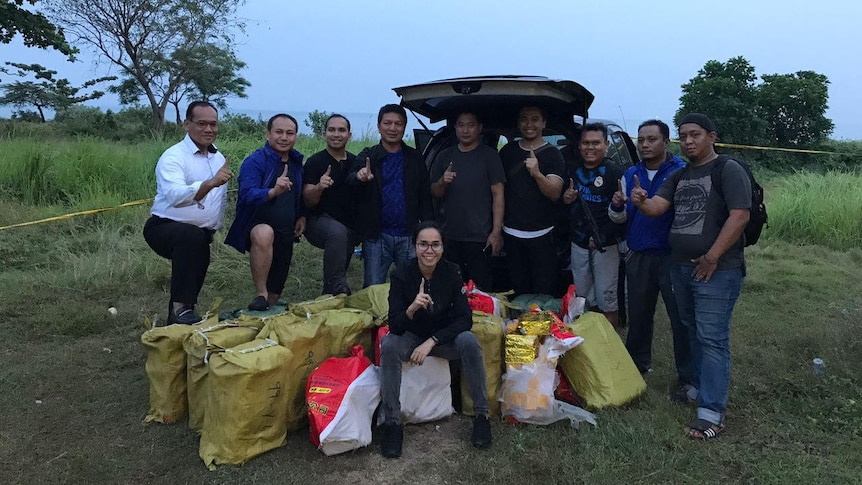 A large group of plain-clothes Indonesian police stand around a pile of bags of drugs.