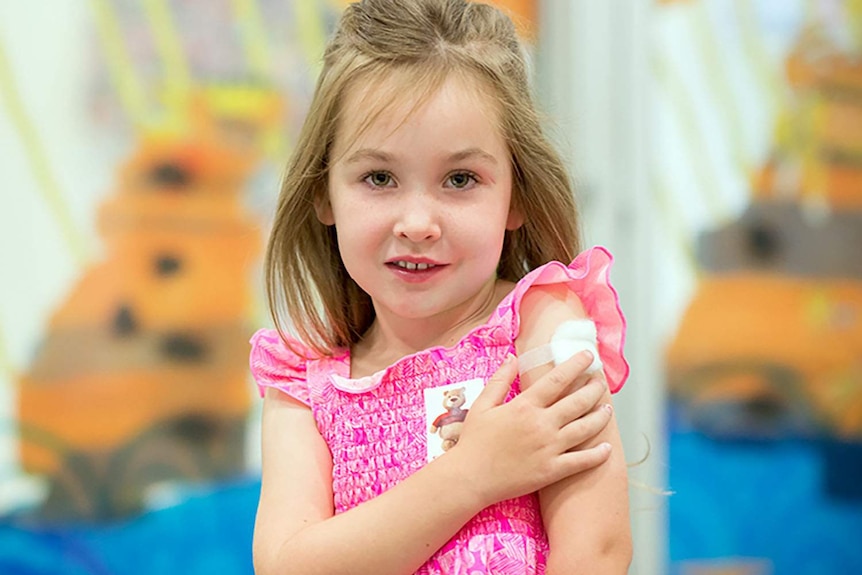 A young girl in pink dress holds her arm with a band-aid after receiving a flu shot.