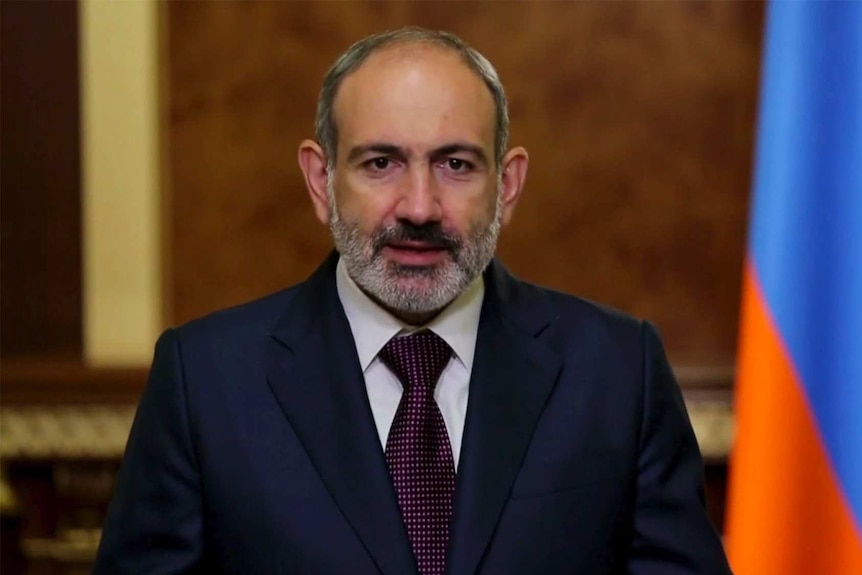 A man in a suit speaking in front of an Armenian flag.