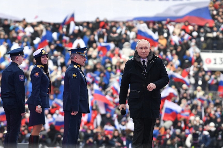 Putin walks towards the camera. Three unfirmed guards watch him go. A blurred stadium crowd makes up the background