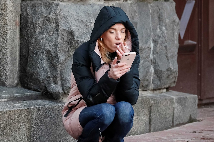 A young woman, wearing a hooded jacket, squats and smokes a cigarette as she looks at her iPhone.