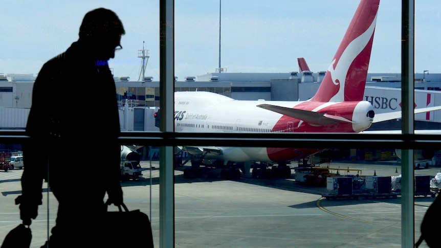 A Qantas plane can be seen on the tarmac at Sydney Airport as a silhouetted man walks past a window inside the terminal.