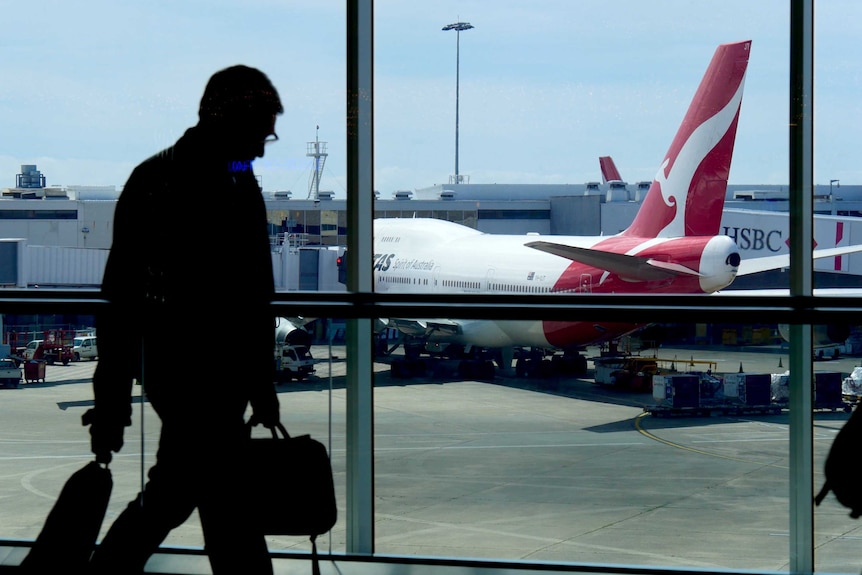A Qantas plane can be seen on the tarmac at Sydney Airport as a silhouetted man walks past a window inside the terminal.