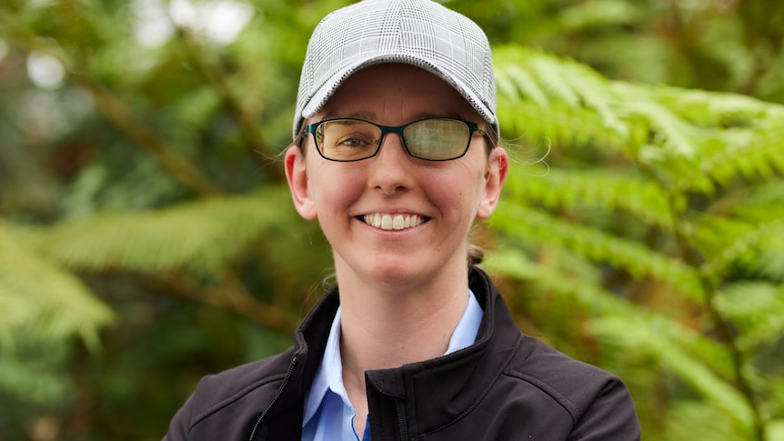 woman in baseball cap, blue shirt and black jacket, with arms folder with green leaves in the background