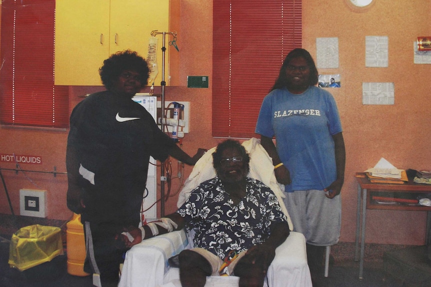 An elderly man sitting in a dialysis clinic surrounded by two children.