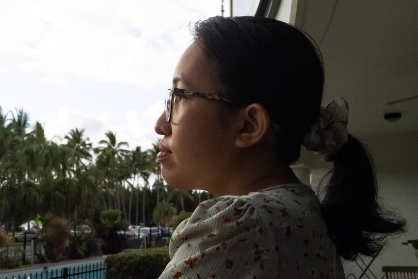 a woman stands on her balcony, overlooking palm trees with a calm expression on her face