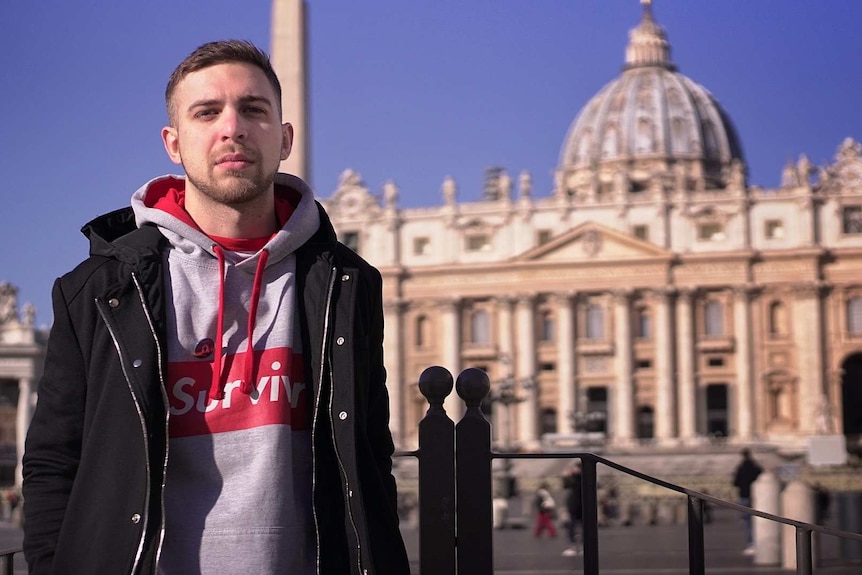 A young man standing in front of St Peter's cathedral wearing a jumper that says 'Survivor'.