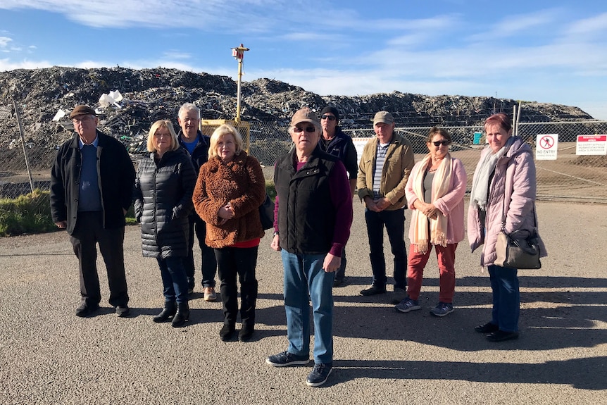 Nine people stand on a road in front of a high mesh fence and towering pile of broken rubbish.