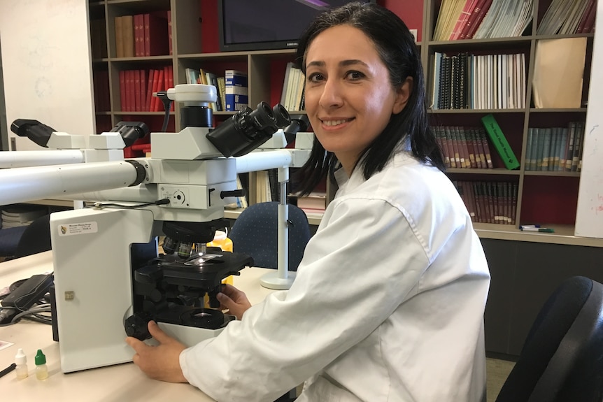 A woman with shoulder-length black hair, wearing a white lab coat, sits at desk with a microscope.