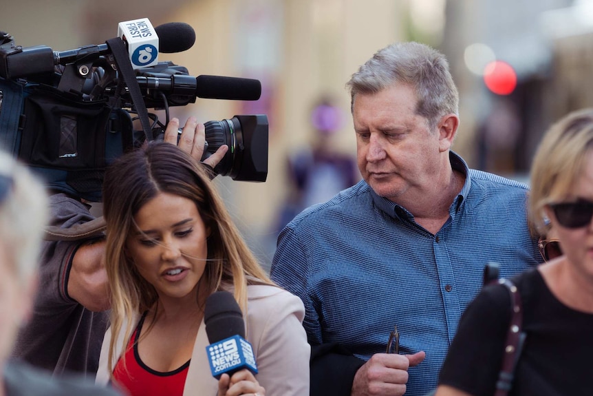 A man in a blue shirt is questioned by reporters as cameras film him on the street.