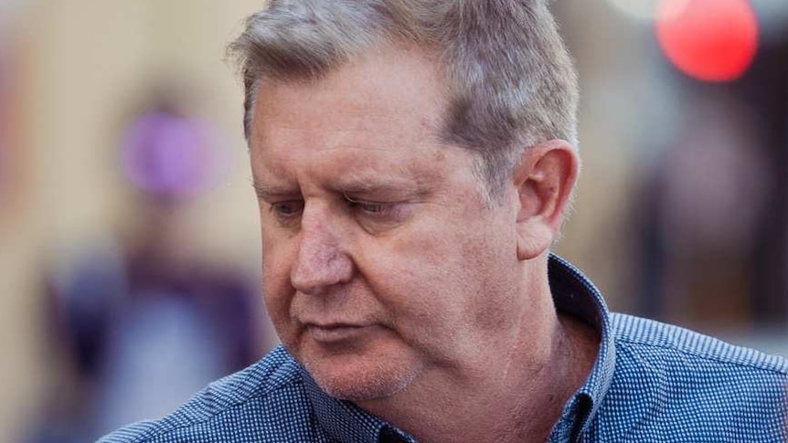 A man in a blue shirt is questioned by reporters as cameras film him on the street.