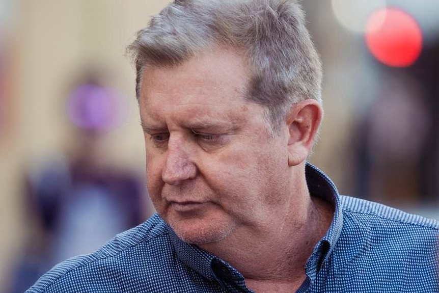 A man in a blue shirt is questioned by reporters as cameras film him on the street.