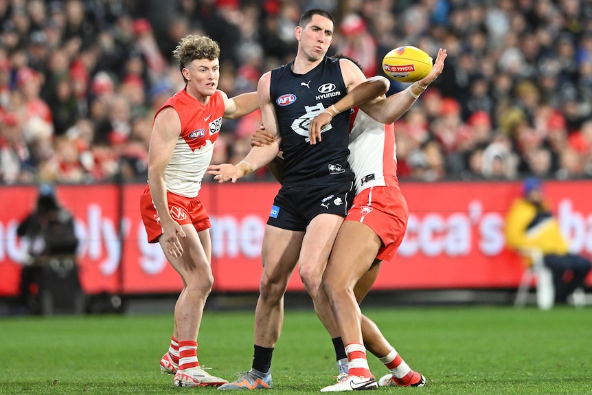 An AFL player in blue, punches the ball away from two opponents in red and white