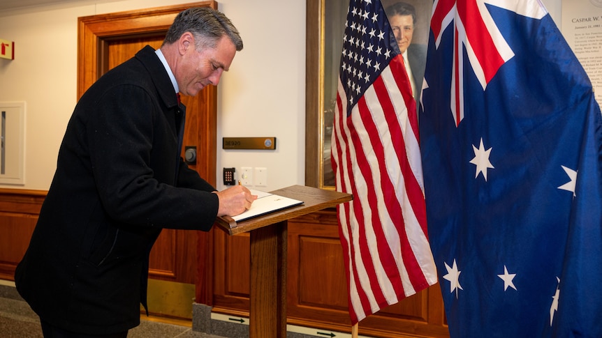 A man signs a book with flags in the background.