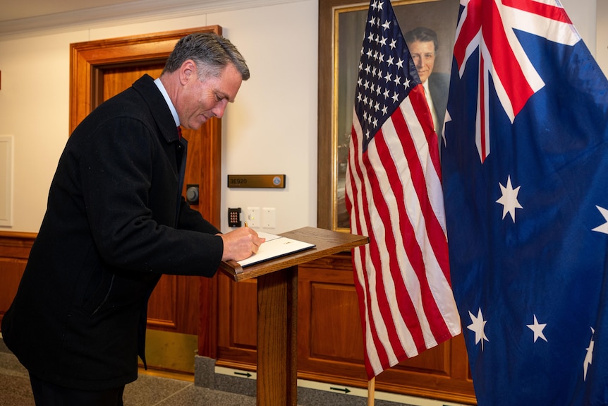 A man signs a book with flags in the background.