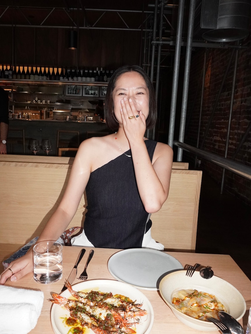 Maggie Zhou smiles, covering her mouth while seated at a restaurant with prawns on the table. Dinner with Jessie Wright