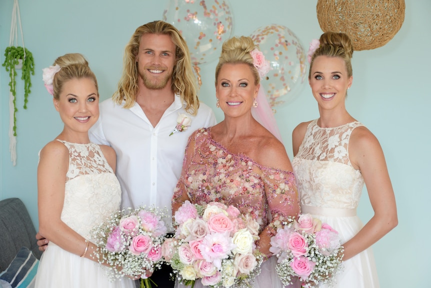 A woman in a blush pink formal dress holds pink and white flowers standing with two adult daughters and adult son. All smiling