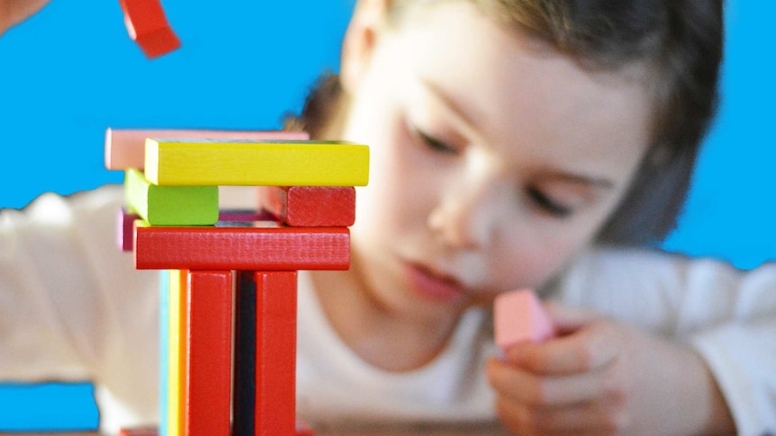 Coloured blocks being stacked by a young girl who's out of focus in the background demonstrating kids learning to lose and win.