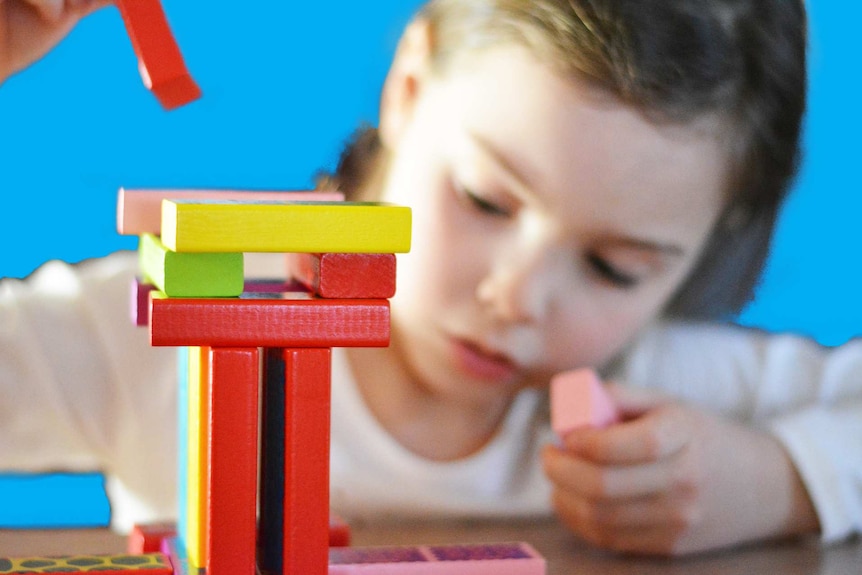 Coloured blocks being stacked by a young girl who's out of focus in the background demonstrating kids learning to lose and win.