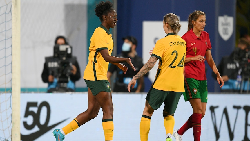 A Matildas striker smiles and reaches out to clap hands with a teammate as they run away from the net after a goal.