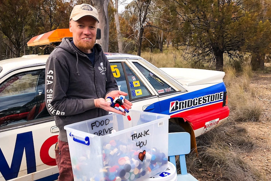 Timothy Miller holding a variety of different coloured bottle caps in front of a car and Australian bushland.