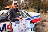Timothy Miller holding a variety of different coloured bottle caps in front of a car and Australian bushland.