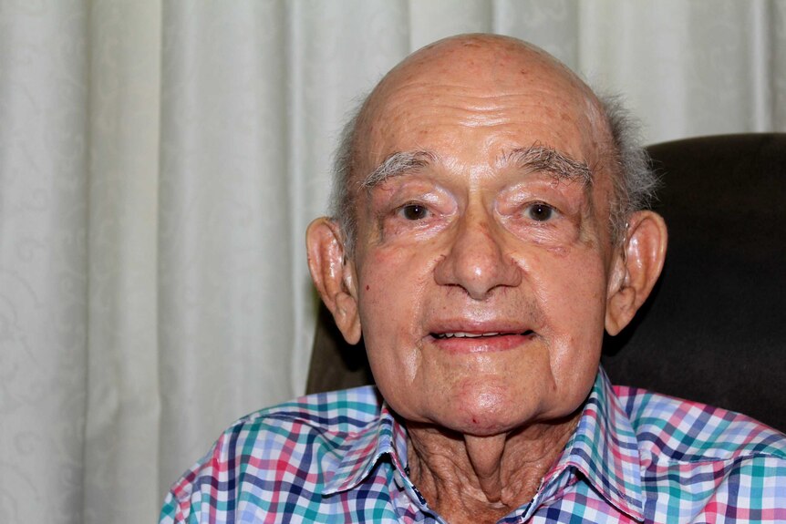 A tight portrait photo of an old man, smiling slightly and wearing a checked button-up shirt.